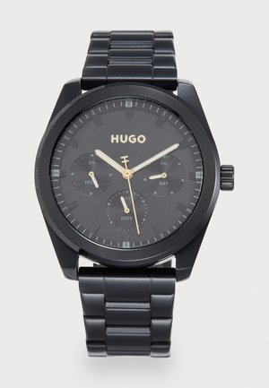 Montre analogique noire avec un boîtier rond, un cadran mat, des accents dorés et trois sous-cadrans pour le jour, la date et l'heure sur 24 heures. Bracelet en maillons métalliques.