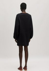Zwarte, geweven jurk met lange, gebogen mouwen die boven de knie valt, gecombineerd met rode sandalen. Eenvoudige en ontspannen silhouet.