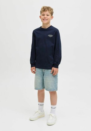 Marineblauw longsleeve shirt met een graphic, lichtblauwe denim shorts, witte sneakers en crew-sokken met zwarte accenten, staand tegen een neutrale achtergrond.