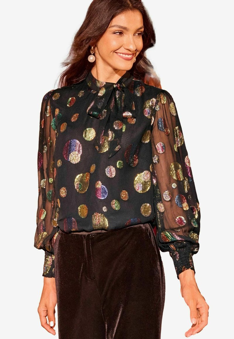 Blusa nera trasparente con pois metallici colorati, maniche lunghe, collo alto con dettagli a fiocco, abbinata a pantaloni di velluto scuro.