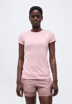 Kobieta z krótkimi włosami, ubrana w jasnoróżową koszulkę z krótkim rękawem i dopasowane szorty, stoi na tle jednolitego białego tła.