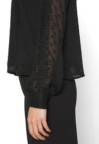 Manche de blouse noire transparente avec texture à pois et bordure en dentelle, resserrée au poignet, montrée sur le bras d'une personne sur un fond uni.