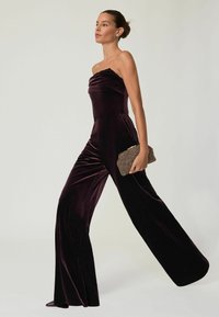 Bársony, szilva színű, pánt nélküli overál széles lábakkal és testhez simuló felsőrésszel. Texturált, arany részletekkel díszített táskával párosítva.