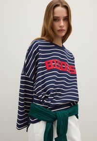 Marineblauw en witte gestreepte blouse met lange mouwen en rode "BISOU" opdruk, gecombineerd met een groene gebreide trui om de taille geknoopt.