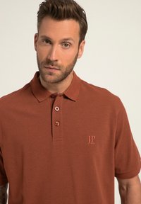 Polo w kolorze rdzawym, wykonany z bawełny, z dekoltem na dwa guziki i małym haftowanym logo po lewej stronie na klatce piersiowej. Krótkie rękawy.