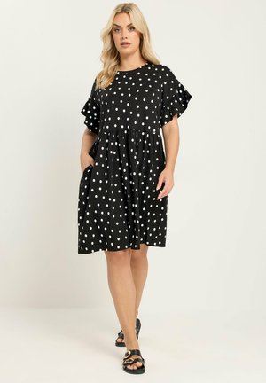 Blonde Frau in schwarzem knielangem Kleid mit weißen Punkten und kurzen Rüschenärmeln, die mit den Händen in den Taschen und schwarzen Sandalen läuft.