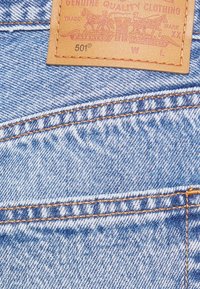 Light blue denim fabric with contrasting orange stitching; features a tan leather patch with embossed branding and model number.