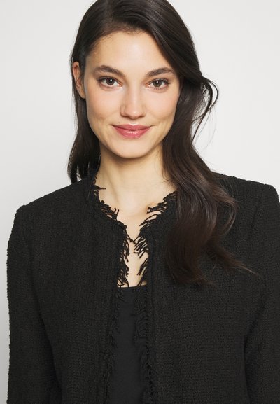 Veste noire texturée avec un col à franges, dotée d'un col rond et d'une conception à ouvert devant. Portée sur un haut noir ajusté.