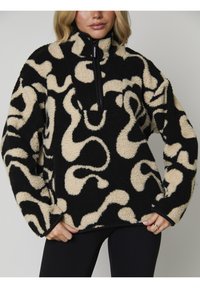 Pull en polaire noir et beige avec un col zippé, présentant un motif tourbillonnant et une surface douce et texturée.