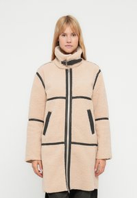 Cappotto in pile beige con accenti neri, colletto alto, chiusura con zip e tasche laterali, caratterizzato da un design strutturato e imbottito.