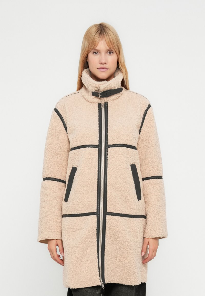 Cappotto in pile beige con accenti neri, colletto alto, chiusura con zip e tasche laterali, caratterizzato da un design strutturato e imbottito.