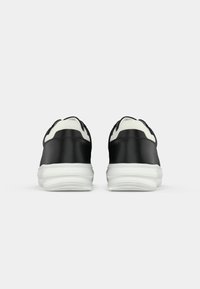 Une paire de baskets noires avec des colliers rembourrés blancs et de grosses semelles blanches, vues de l'arrière sur un fond blanc uni.