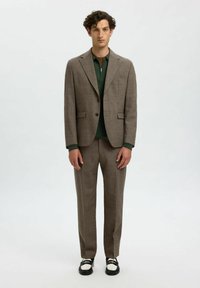 Traje marrón con solapa muesca, una camiseta verde con cremallera debajo y zapatos negros con acentos blancos. Corte ajustado, textura suave.
