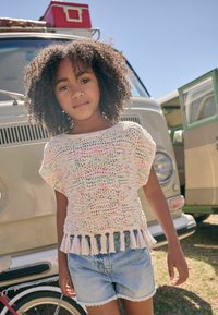 Gehaakte top met een multicolor patroon en een franjeszoom, gecombineerd met een lichte denimshort. De achtergrond bevat vintage voertuigen.
