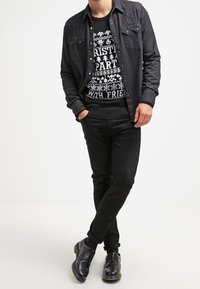 Camicia nera a pulsante sovrapposta a un maglione di Natale nero con testo grafico bianco e motivi, abbinata a pantaloni e scarpe nere.