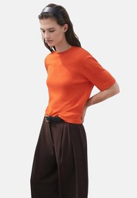 Oranges, gestricktes T-Shirt mit kurzen Ärmeln und Rundhals-Ausschnitt, kombiniert mit dunkelbraunen, gefaltenen Hosen und einem schwarzen Gürtel, der eine kontrastreiche Farbkombination schafft.