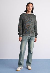 Young man with shoulder-length hair wearing a green textured sweater, faded blue jeans, and white sneakers standing on a blue floor.