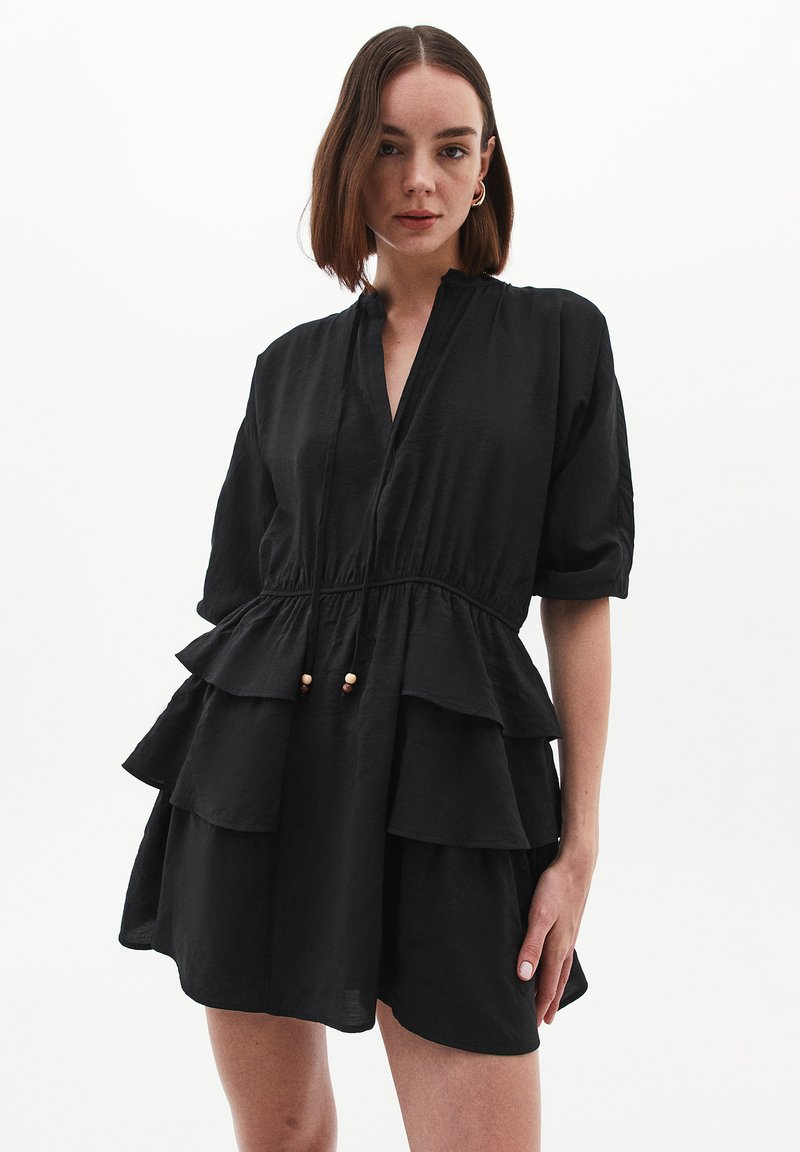 Schwarzes Kleid mit V-Ausschnitt, kurzen Ärmeln und geschichteten Rüschen. Hat eine taillierte Taille und dekorative Holzperlen an verstellbaren Bindebändern.