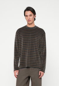 Νέος άνδρας που στέκεται μπροστά σε λευκό φόντο, φορώντας ένα μαύρο και καφέ καρό πουκάμισο με μακριά μανίκια και καφέ παντελόνι.