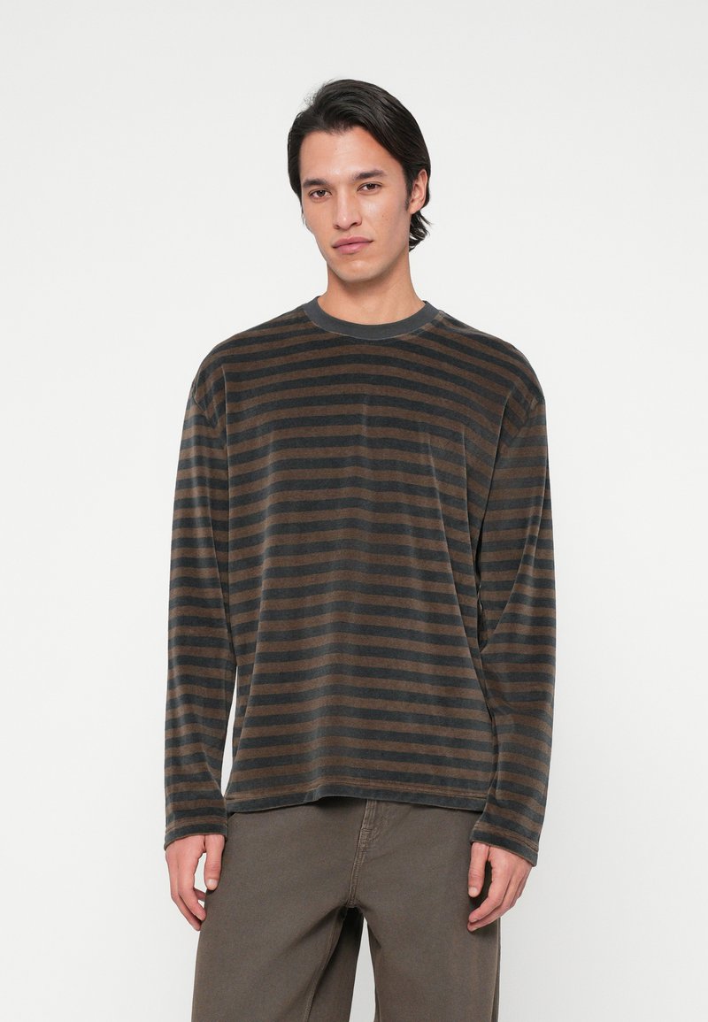 Νέος άνδρας που στέκεται μπροστά σε λευκό φόντο, φορώντας ένα μαύρο και καφέ καρό πουκάμισο με μακριά μανίκια και καφέ παντελόνι.