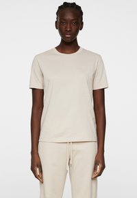 Beige bomullströja med rund halsringning, korta ärmar och en diskret logotyp på bröstet. Tyget verkar slätt och lätt.