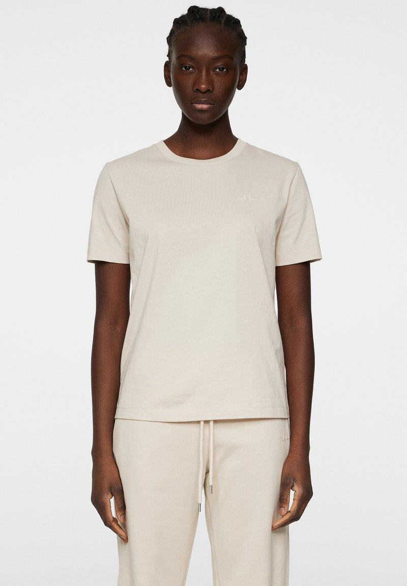 Beige bomullströja med rund halsringning, korta ärmar och en diskret logotyp på bröstet. Tyget verkar slätt och lätt.