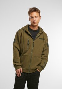 Felpa con zip di colore verde oliva, con cappuccio regolabile con coulisse, tasche con zip e patch in velcro sulle maniche. Tessuto morbido e vestibilità rilassata.