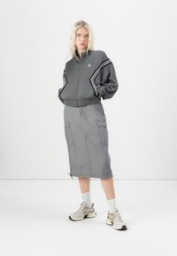 Veste grise à fermeture éclair avec des rayures noires sur les côtés et un col montant, associée à une jupe grise de longueur genou avec des poches cargo. Des baskets beiges complètent le look.