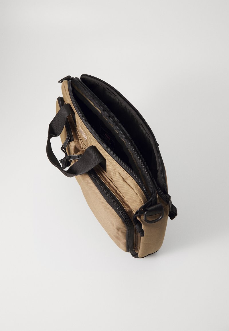 Beige Laptop-Tasche mit schwarzen Griffen und mehreren Reißverschlussfächern, geöffnet vor einem schlichten weißen Hintergrund.