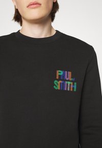 Personne portant un sweat-shirt noir à col ras du cou avec "PAUL SMITH" coloré brodé sur le côté gauche de la poitrine.