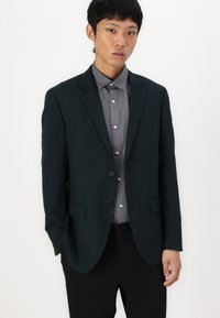 Dunkelgrüner Blazer mit tailliertem Schnitt, zwei Knöpfen, Reverskragen und vorderen Taschen über einem hellgrauen Hemd mit Knöpfen. Glatte Textur.