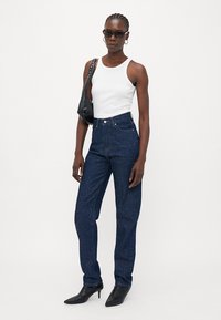 Hoge taille donkerdenim jeans met gespikkelde accenten, gecombineerd met een wit tanktop en een zwarte handtas, aangevuld met elegante zwarte haklaarzen.