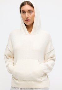 Cream hooded sweatshirt in soft fabric, featuring a front pocket and ribbed hem. Subtle grey accents along the sleeves. Relaxed fit.