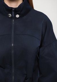 Mørkeblå puffjakke med høy krage, glidelås og sølvfargede trykknapper. Stoffet ser glatt ut med en lett glans.