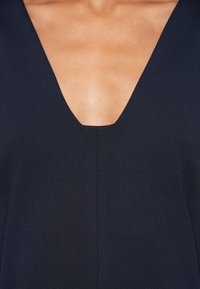 Artimas asmens vaizdas, vilkinčio tamsiai mėlyną, V formos kaklo drabužį, kuriame matyti raktikaulis ir viršutinė krūtinės dalis su centre esančia siūle.