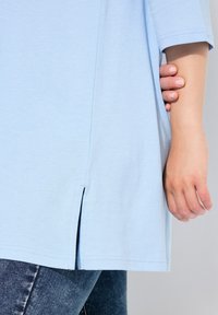 Top azzurro chiaro a maniche corte in tessuto morbido, con fessure laterali all'orlo, indossato sopra jeans blu scuro. Texture liscia e vestibilità comoda.