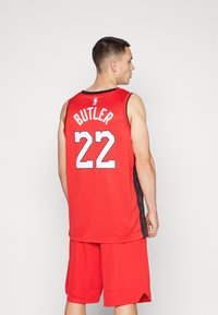 Camiseta de baloncesto roja con acentos negros, diseño sin mangas, que presenta "BUTLER" y el número "22" en la parte posterior. La camiseta tiene una superficie texturizada.