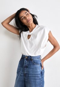 Witte top met korte mouwen, voorzien van een keyhole-detail en een knoopaccent, gecombineerd met hoog getailleerde blauwe denim jeans met een knoopsluiting.