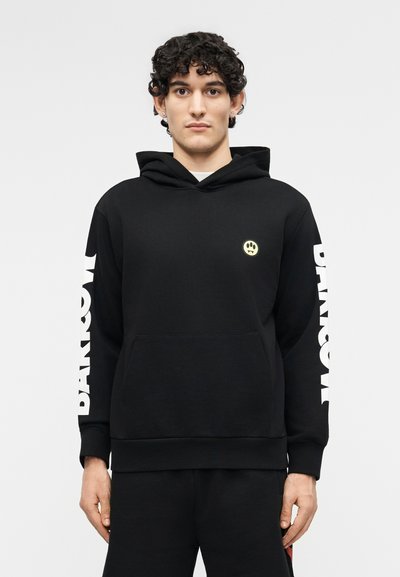 Ung mann med krøllete hår iført en svart hettegenser med hvit "DAMNSON"-tekst på ermene og et lite gult smilefjes på brystet.