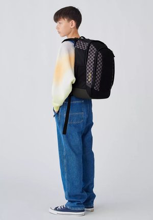 Jeune personne portant un sac à dos noir à carreaux, un jean décontracté, des baskets et un pull léger dégradé, debout de profil sur un fond uni.