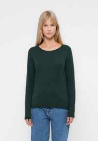 Pull en maille verte avec un col rond, des manches longues et des poignets côtelés, associé à un jean en denim bleu clair.
