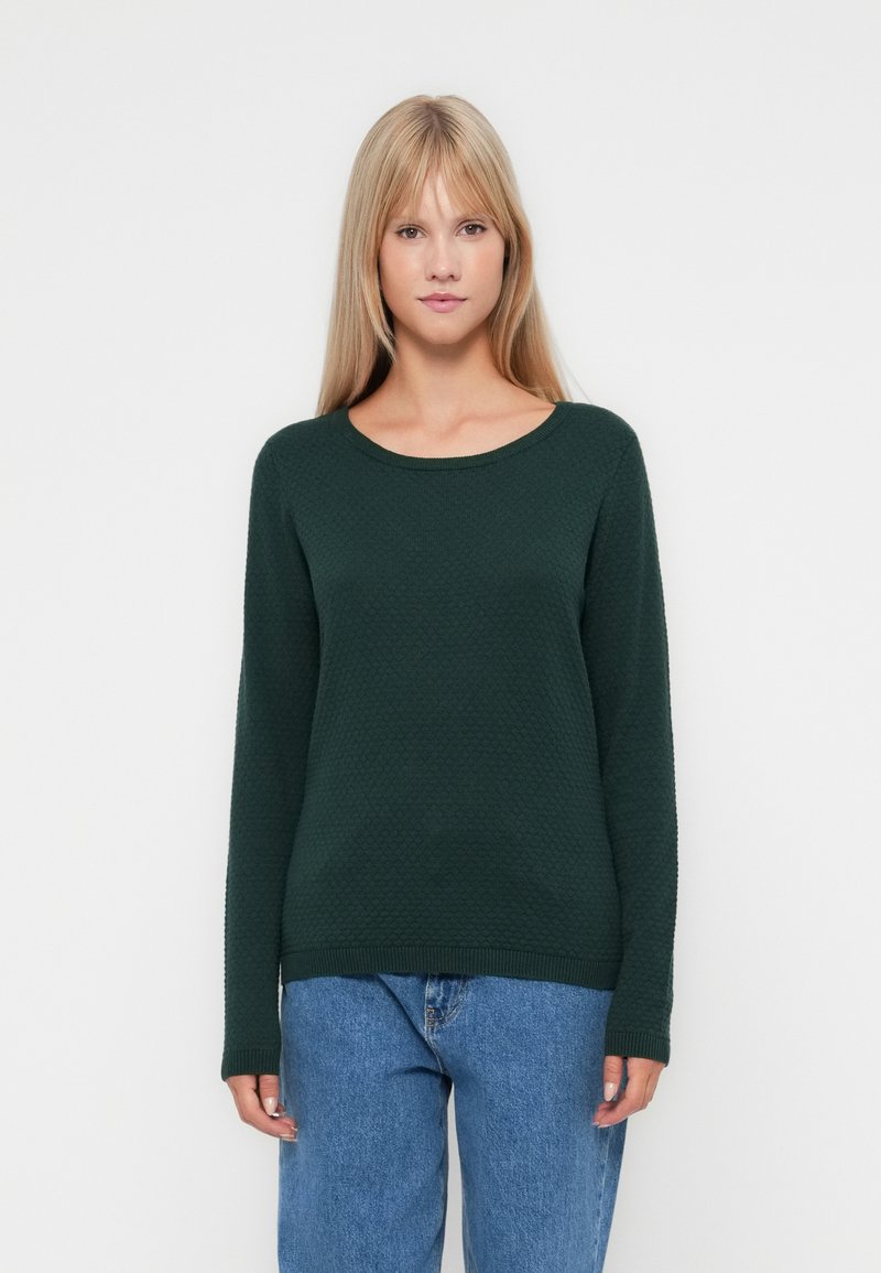 Pull en maille verte avec un col rond, des manches longues et des poignets côtelés, associé à un jean en denim bleu clair.