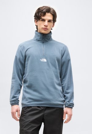 Mladý muž oblečený v modrom fleece pulóvri so zipsom do polovice a logom „The North Face“ a čiernych nohaviciach, stojaci pred jednoduchým svetlým pozadím.