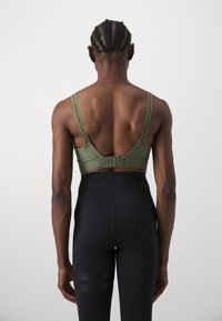 Sutiã desportivo verde-oliva com alças ajustáveis, fecho de gancho e tecido texturizado. Combinado com leggings ajustadas pretas com riscas.