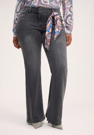 Frau trägt hoch taillierte, ausgewaschene schwarze Jeans mit einem fließenden Paisley-Schal, der an der Gürtelschlaufe gebunden ist, und spitze graue Schuhe, die darunter sichtbar sind.