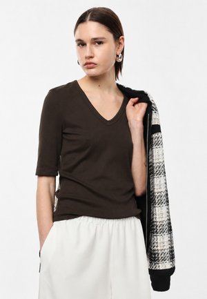 Donna con capelli corti castani che indossa una maglietta scura con scollo a V e pantaloni bianchi, tenendo una giacca a quadri bianca e nera sulla spalla.