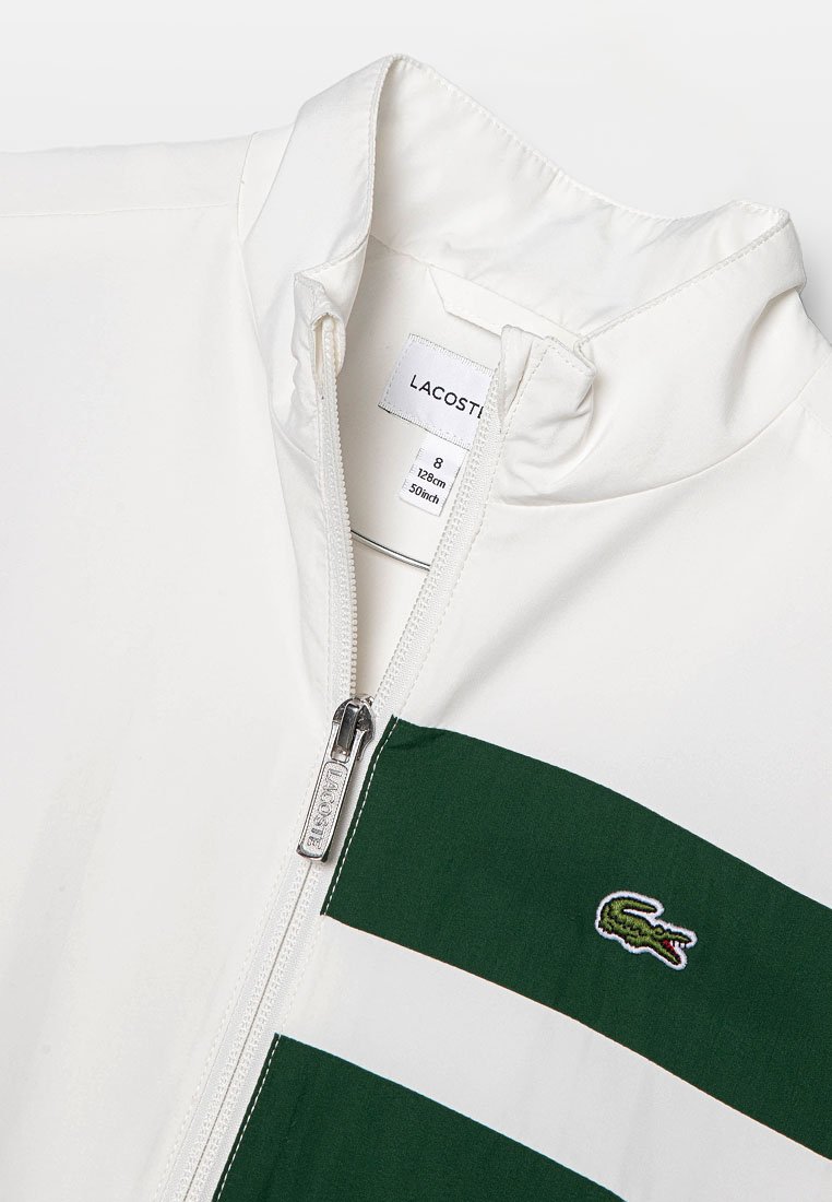 Veste blanche zippée avec des rayures vertes, fabriquée en tissu lisse. Elle possède un col montant et un logo de crocodile brodé sur la poitrine.