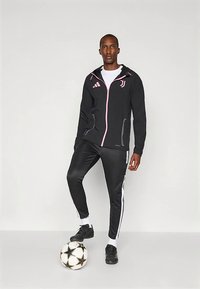 Chaqueta deportiva negra con detalles en rosa y logo de Adidas, combinada con pantalones negros ajustados con rayas blancas, de pie junto a un balón de fútbol.