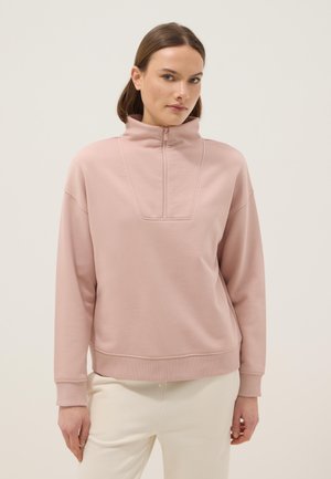 Frau mit braunen Haaren trägt ein hellrosa Reißverschluss-Sweatshirt mit hohem Kragen und cremeweiße Hose, steht vor einem schlichten hellen Hintergrund.