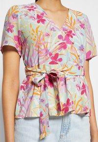 Blouse florale avec un décolleté en V, des manches courtes et une ceinture à nouer. Le tissu est léger avec une base bleu pastel et des motifs vibrants de rose, orange et vert.